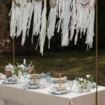 Boho-stiilis piknikulaud õues - pikniku laud, kreemika lina, helesiniste klaaside ja salvrättidega. Taustal rippuvad heledad unenäopüüdjad, mis loovad õhulise ja romantilise meeleolu. Just See rent pikniku lauakatte komplekt vabas õhus pidudeks või tubaseks piknikuks.