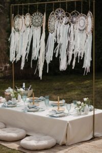 Boho-stiilis piknikulaud õues - pikniku laud, kreemika lina, helesiniste klaaside ja salvrättidega. Taustal rippuvad heledad unenäopüüdjad, mis loovad õhulise ja romantilise meeleolu. Just See rent pikniku lauakatte komplekt vabas õhus pidudeks või tubaseks piknikuks.