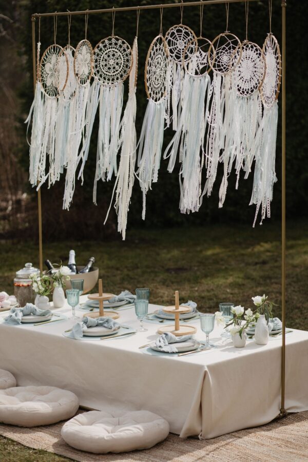 Boho-stiilis piknikulaud õues - pikniku laud, kreemika lina, helesiniste klaaside ja salvrättidega. Taustal rippuvad heledad unenäopüüdjad, mis loovad õhulise ja romantilise meeleolu. Just See rent pikniku lauakatte komplekt vabas õhus pidudeks või tubaseks piknikuks.
